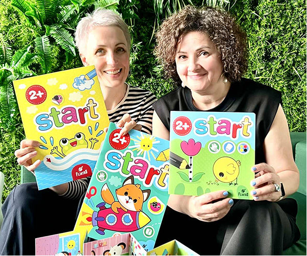 Two smiling women hold colorful children’s activity books titled Start 2+ in front of a green plant wall.